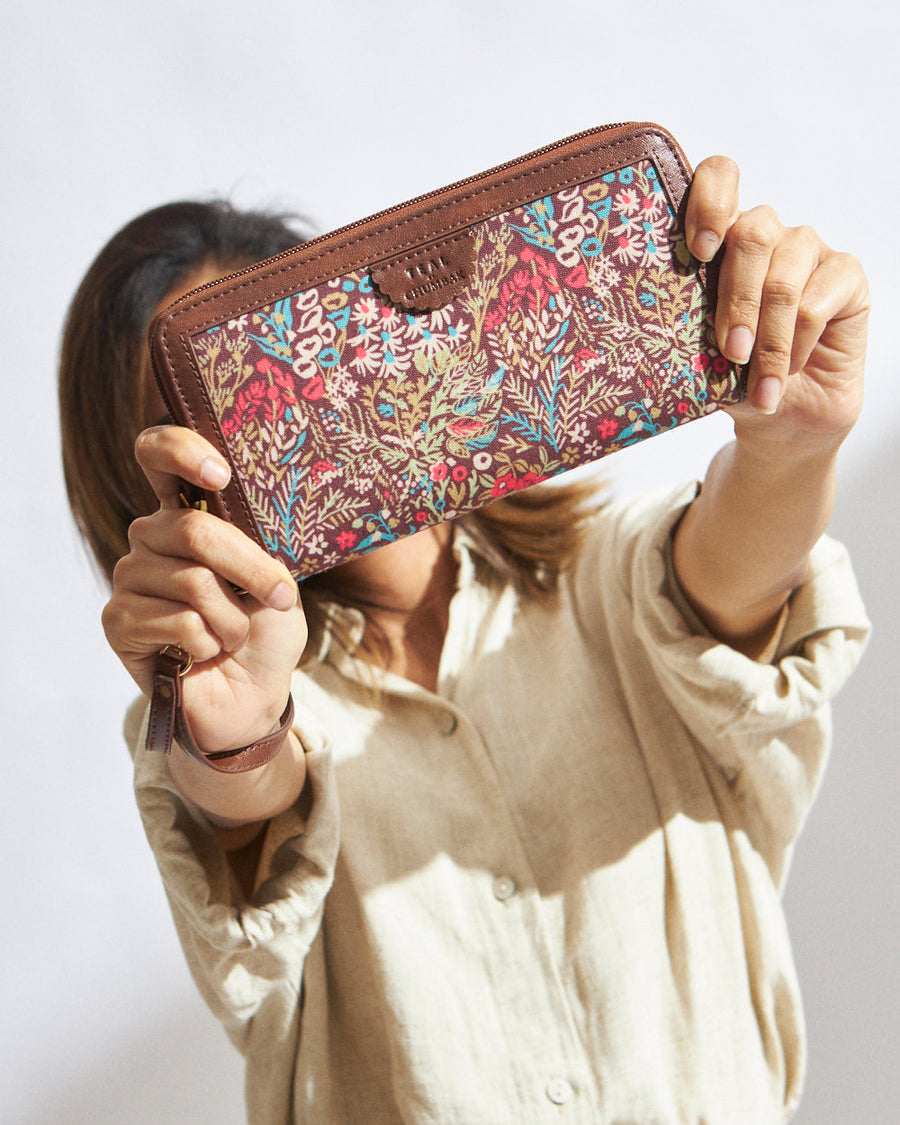 Woman holding a brown floral-patterned long wallet with zipper and wrist strap in front of her face