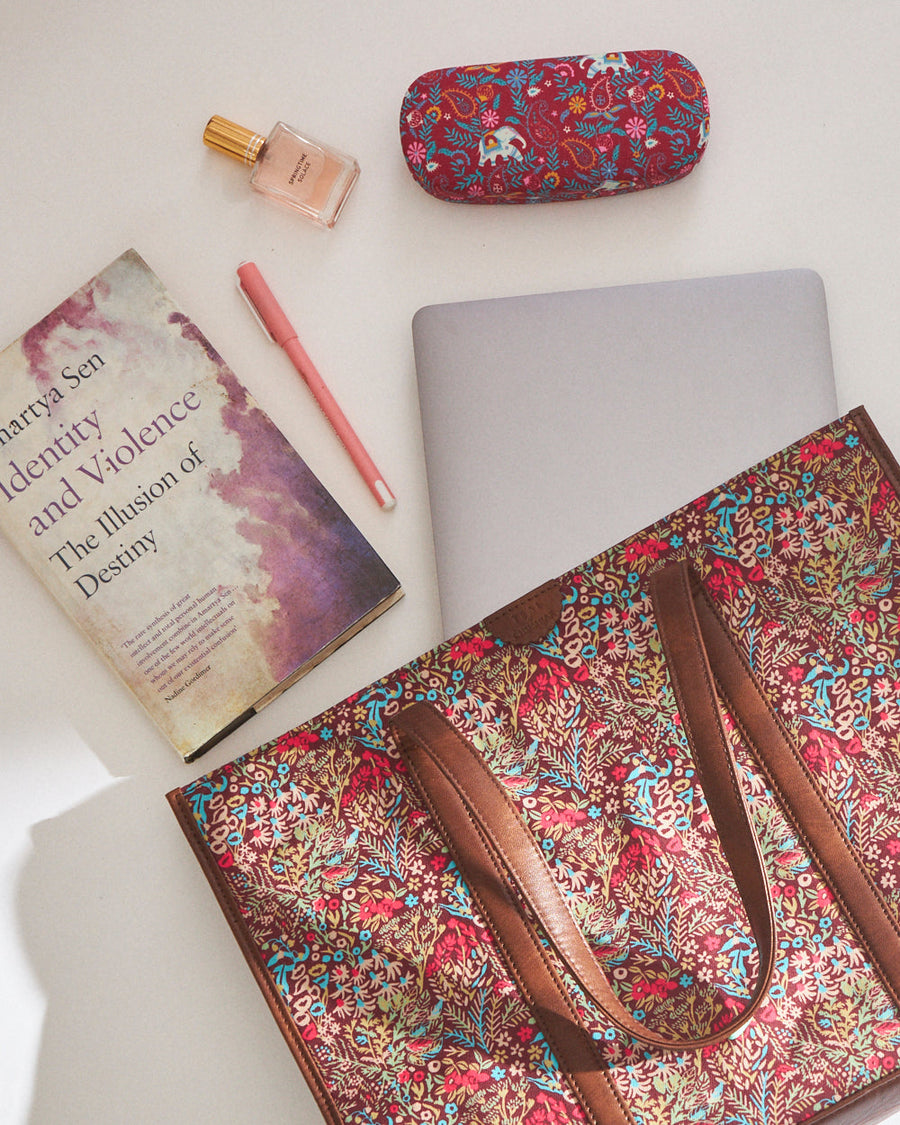 Floral canvas tote bag partially open showing large internal compartment with laptop, book, cosmetic bottle, pen, and floral eyeglass case