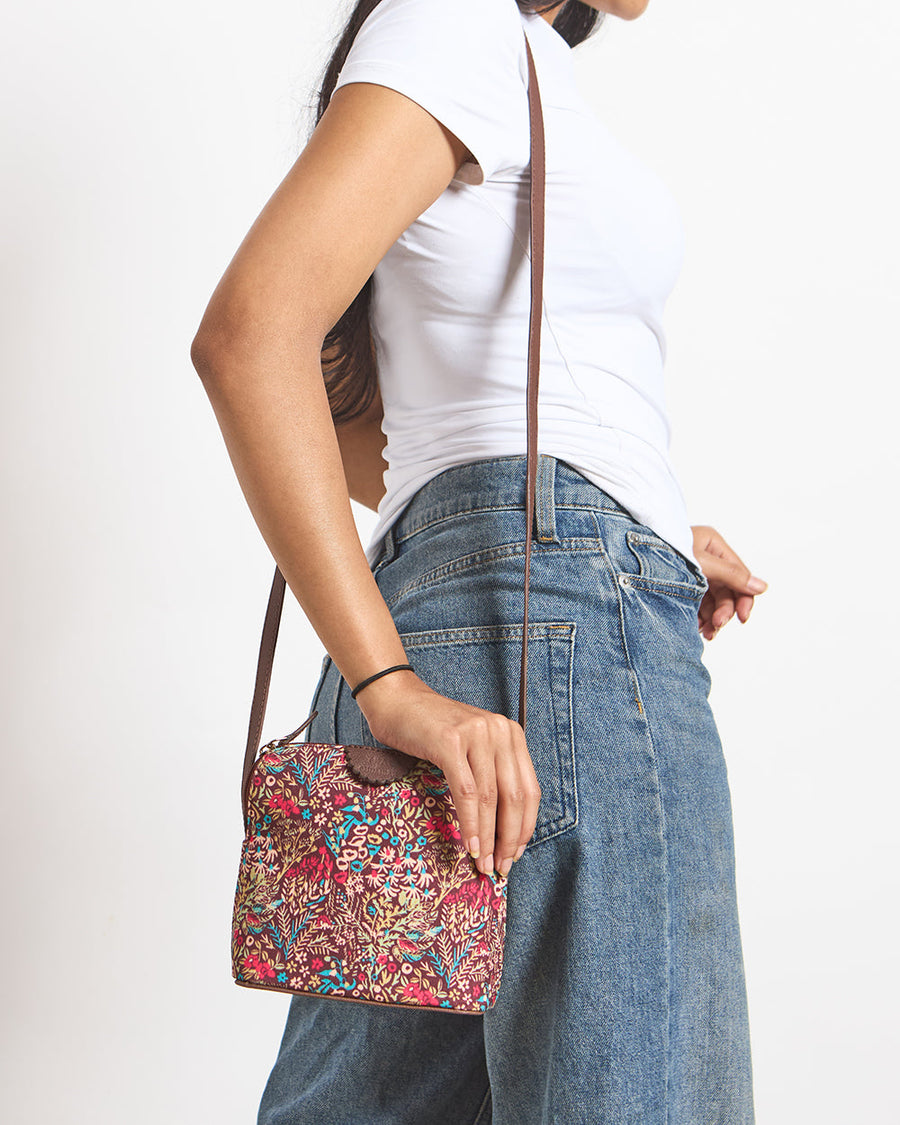 Woman wearing a white t-shirt and blue jeans carrying a compact Rainforest Sling Bag with a colorful botanical print and brown adjustable strap.