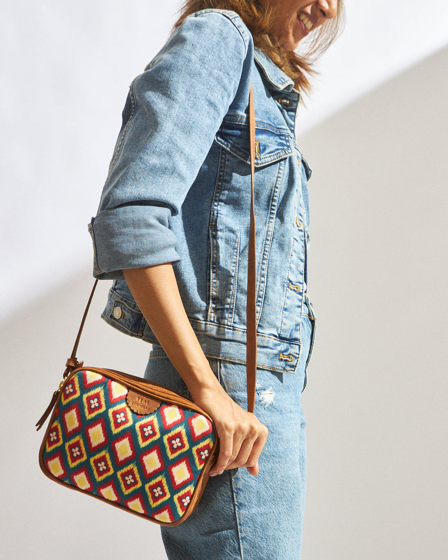 Woman wearing denim jacket and jeans carrying a rectangular sling bag with colorful ikkat geometric pattern and brown adjustable faux leather strap