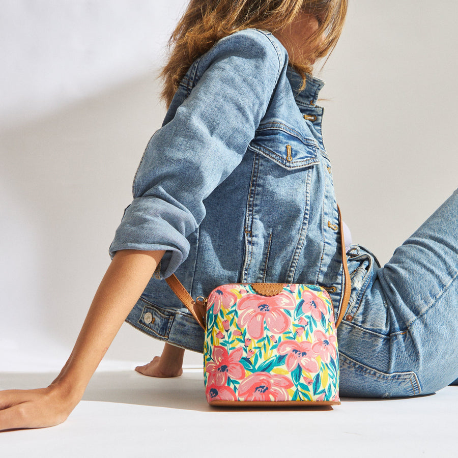 Woman in denim outfit sitting on the floor with a vibrant floral print crossbody bag with tan strap beside her.