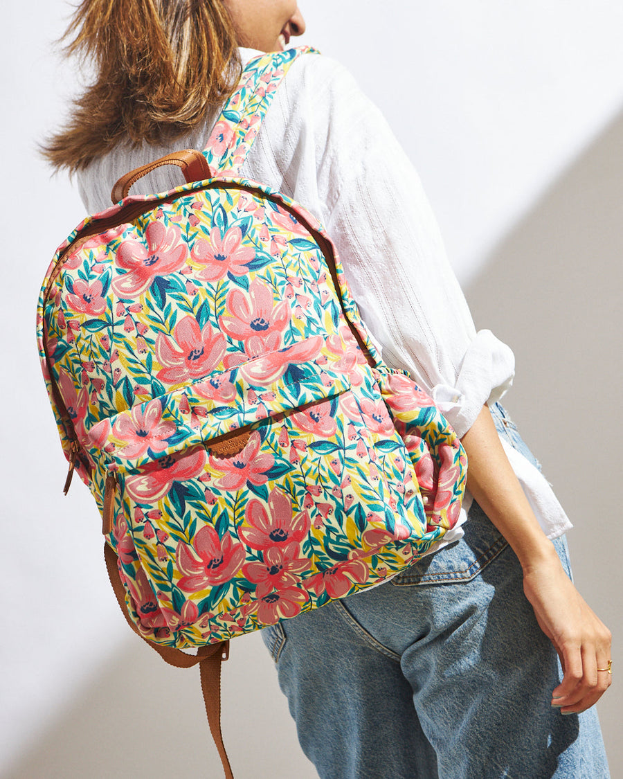 Woman wearing a colorful floral canvas backpack with coral flowers and green leaves on a yellow background, paired with a white shirt and denim jeans.