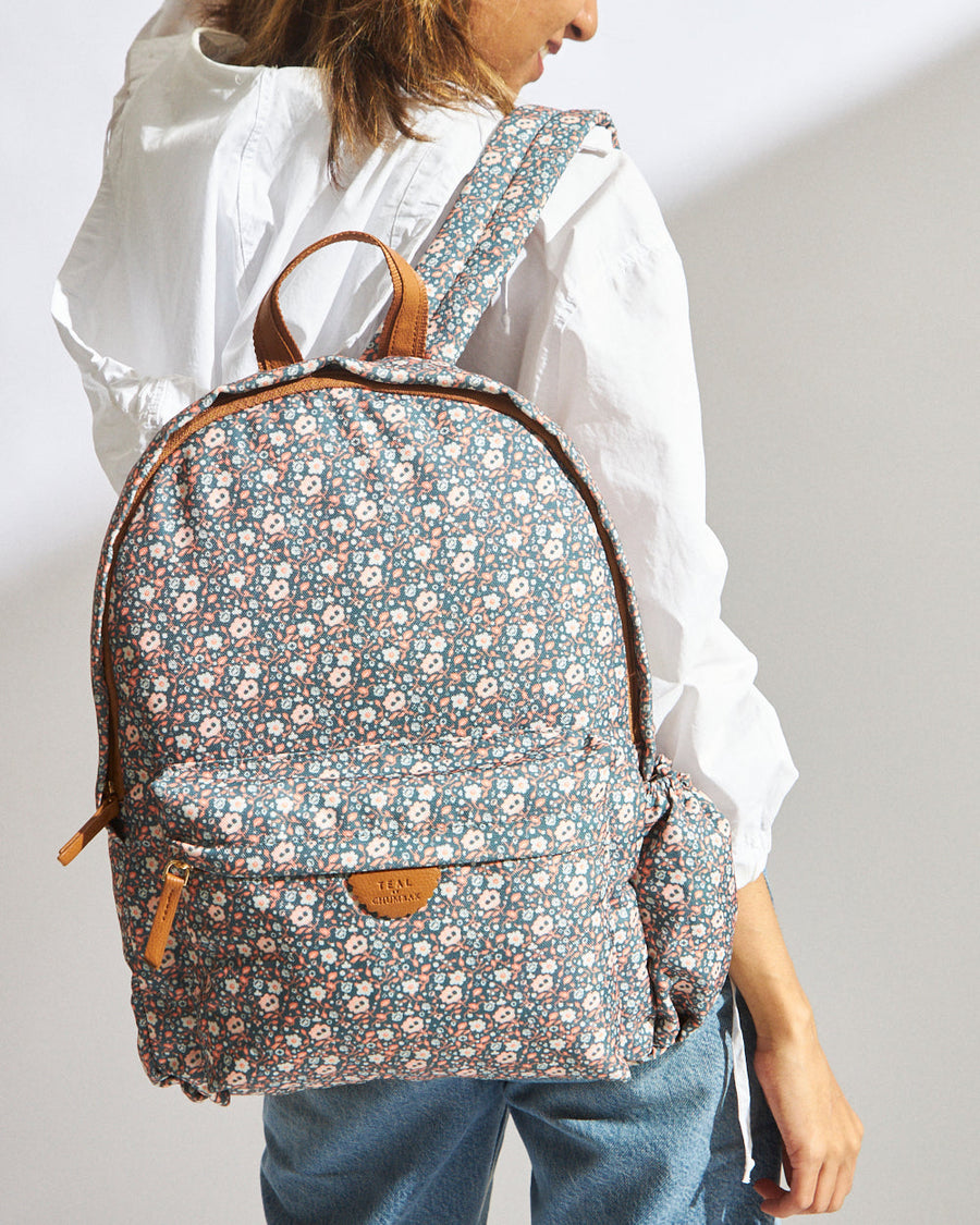 Woman wearing a teal floral patterned backpack with white and peach flowers, featuring padded shoulder straps and multiple pockets
