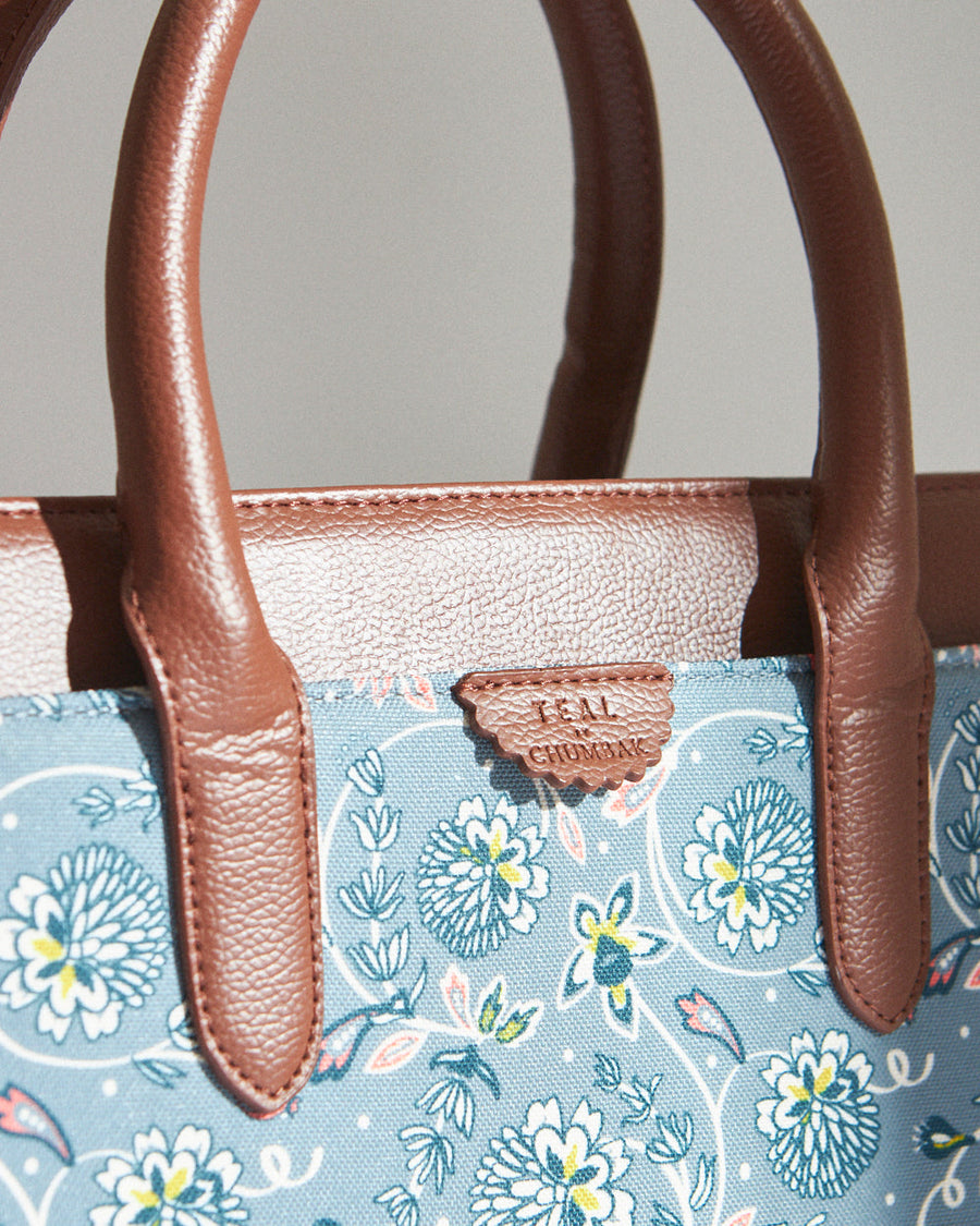 Close-up of handbag's brown vegan leather handles and teal floral canvas material highlighting texture and brand name plate