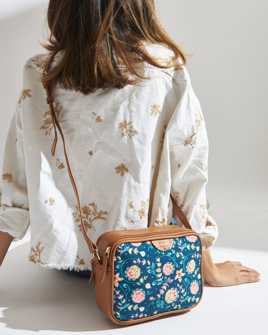 Woman wearing a floral print box sling bag with teal fabric and tan leather accents, shown from the back with strap over shoulder.