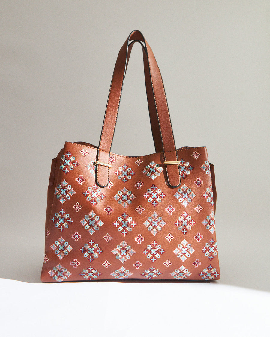 Front view of a tan tote bag showcasing symmetrical cross stitch embroidery and gold-tone strap buckles