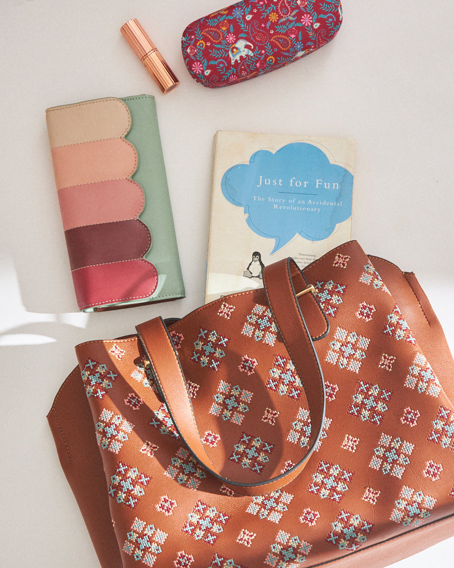 Top view of a tan cross stitch tote bag surrounded by accessories including a wallet and lipstick