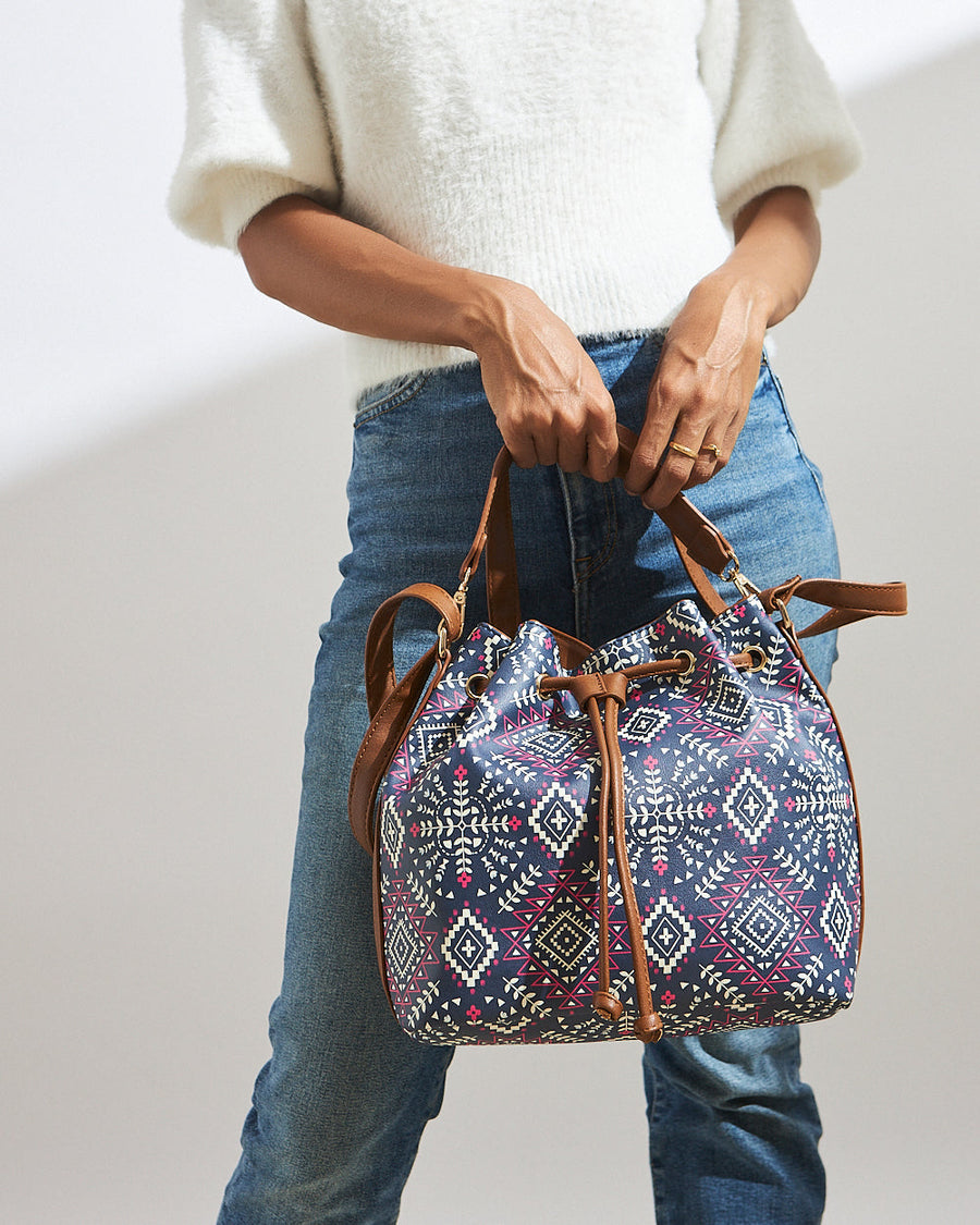 Woman holding navy blue bucket bag with Aztec geometric print, brown leather handles and drawstring closure, styled with white sweater and denim jeans