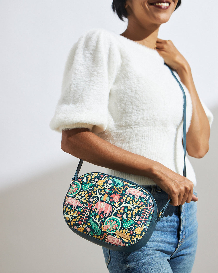 Woman wearing a white textured top and blue jeans carrying a Sundarban-inspired embroidered sling bag with colorful jungle animal motifs and an adjustable strap.
