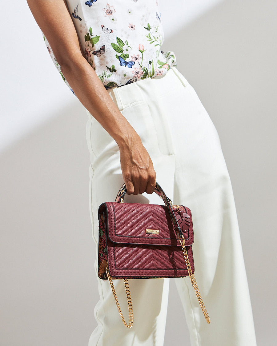 Woman holding a compact quilted burgundy handbag with a gold chain and top handle, paired with white pants and a floral blouse.