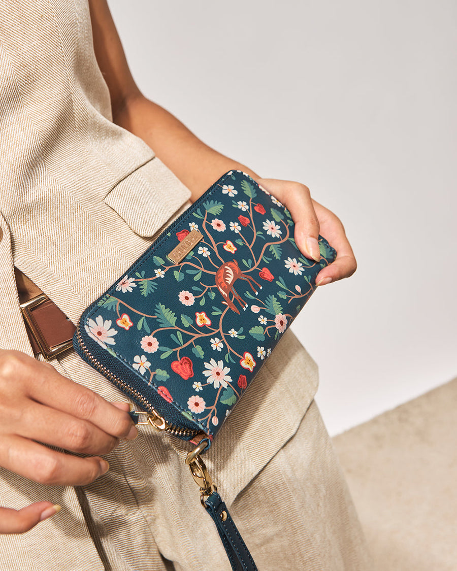 Close-up of a woman's hand holding a teal leather wallet with floral embroidery and a wrist strap, wearing beige textured pants.