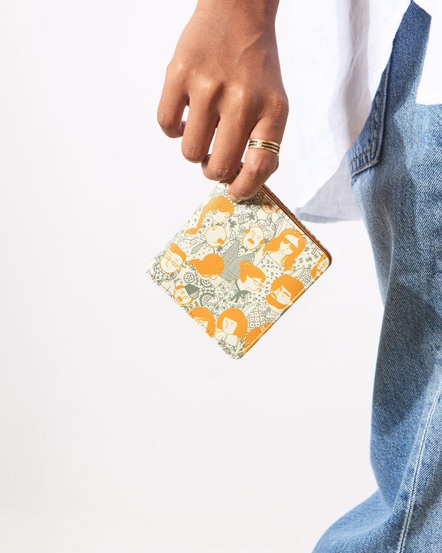 Woman holding a compact mini wallet with a colorful illustrated face pattern in orange and green against a cream background, wearing casual denim and white shirt.