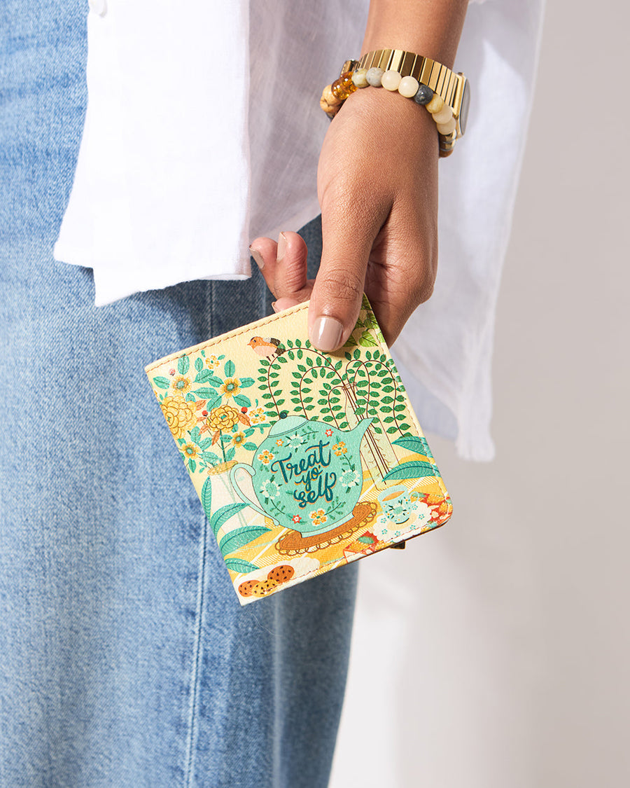 Woman holding a compact mini wallet featuring a turquoise teapot with 'Treat Your Self' text amidst vibrant floral and tea-time illustrations