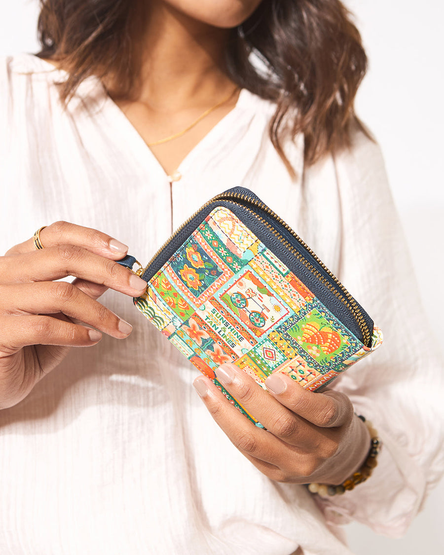 Woman holding a colorful mosaic patterned mini wallet with zipper, wearing a light white blouse and gold jewelry