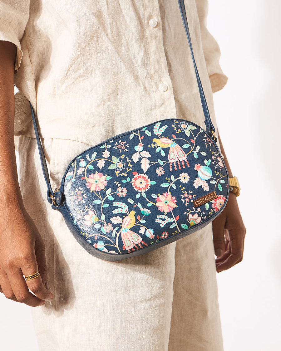 Woman wearing a navy blue floral print crossbody bag with colorful birds, styled with a beige linen outfit