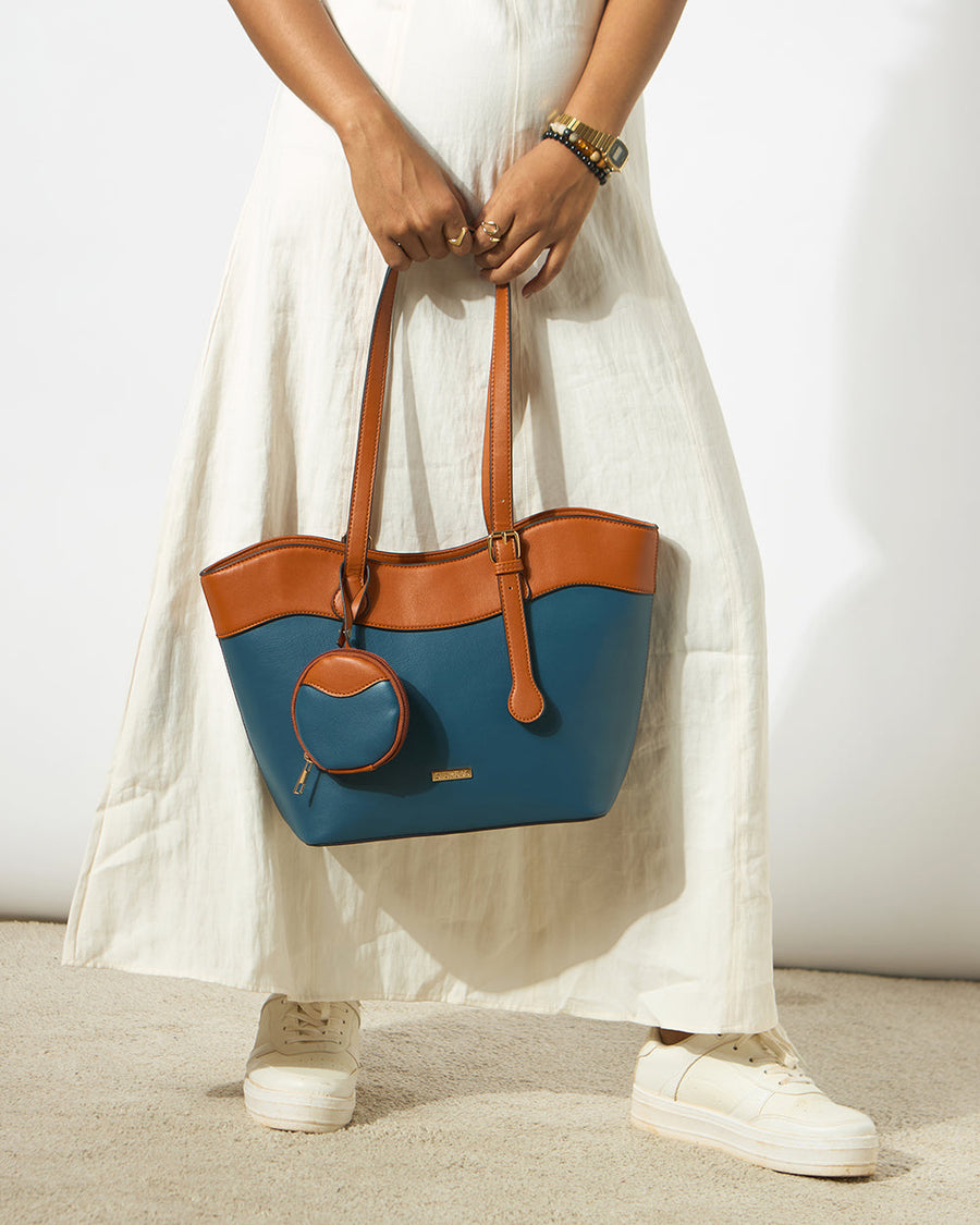 Woman holding a two-tone tote bag in deep teal and caramel brown with a detachable round pouch, paired with a white dress and white sneakers.