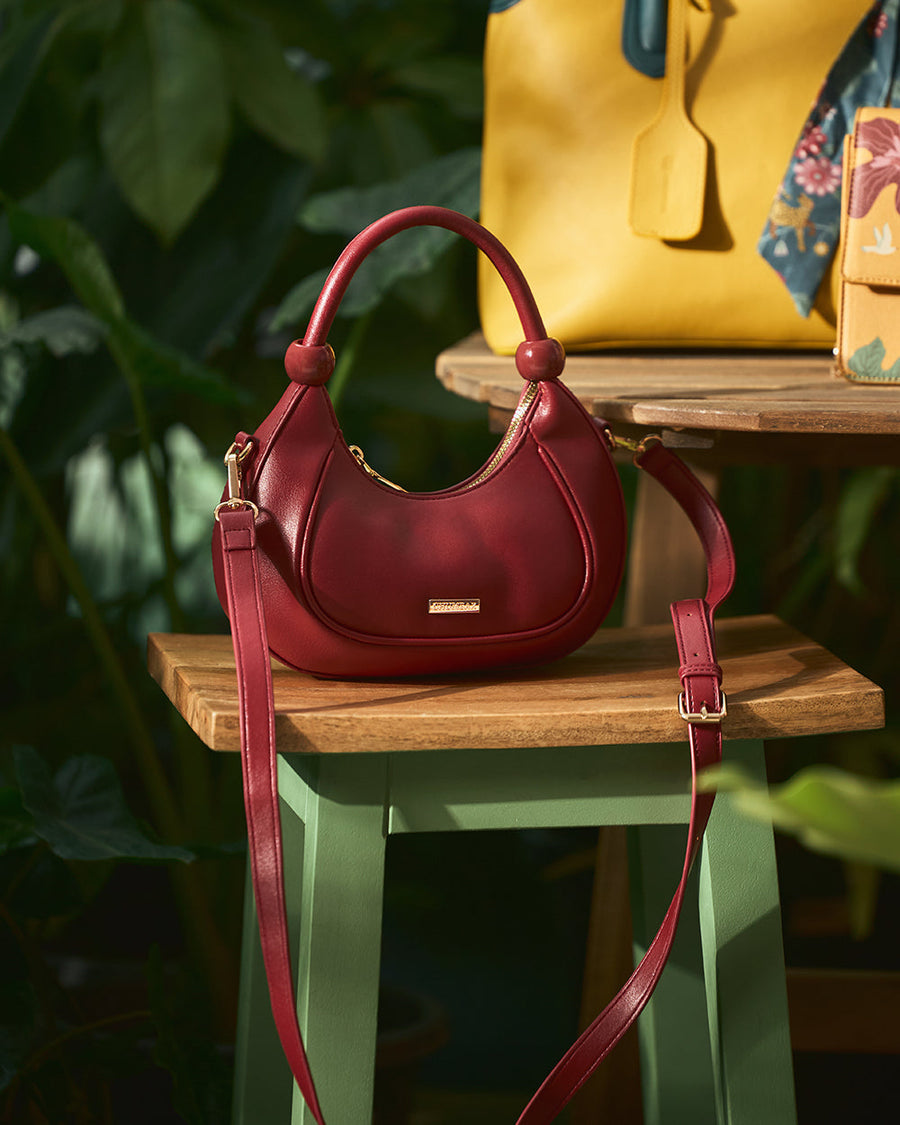 Maroon crescent-shaped vegan leather handbag resting on a wooden stool, featuring a rounded handle and long adjustable strap.