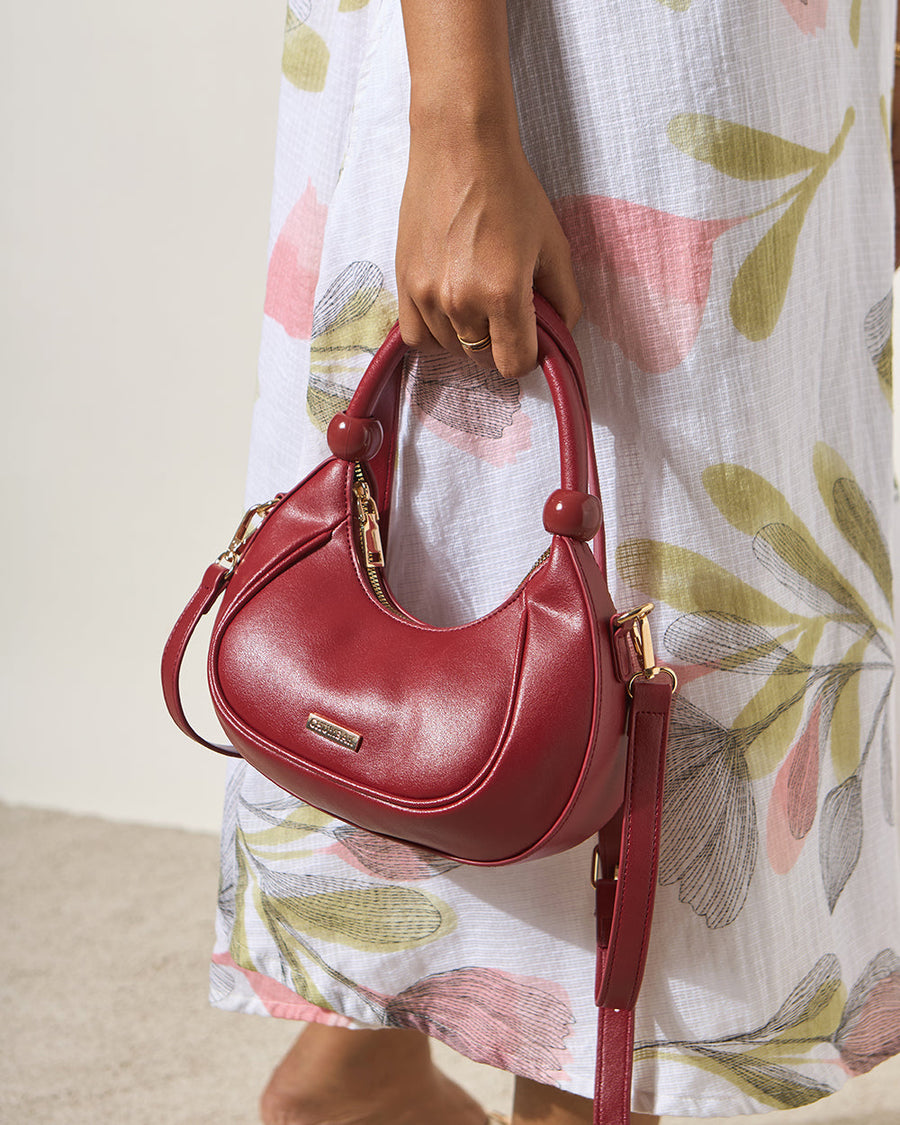 Woman holding the maroon vegan leather crescent handbag by the top handle, paired with a floral patterned dress.