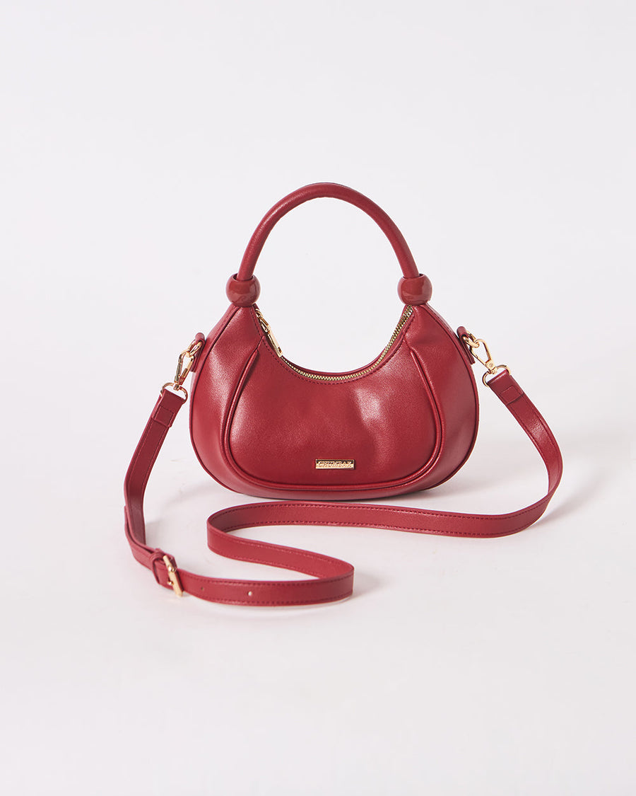 Studio shot of maroon vegan leather crescent handbag with dual handles and adjustable strap on plain white background.