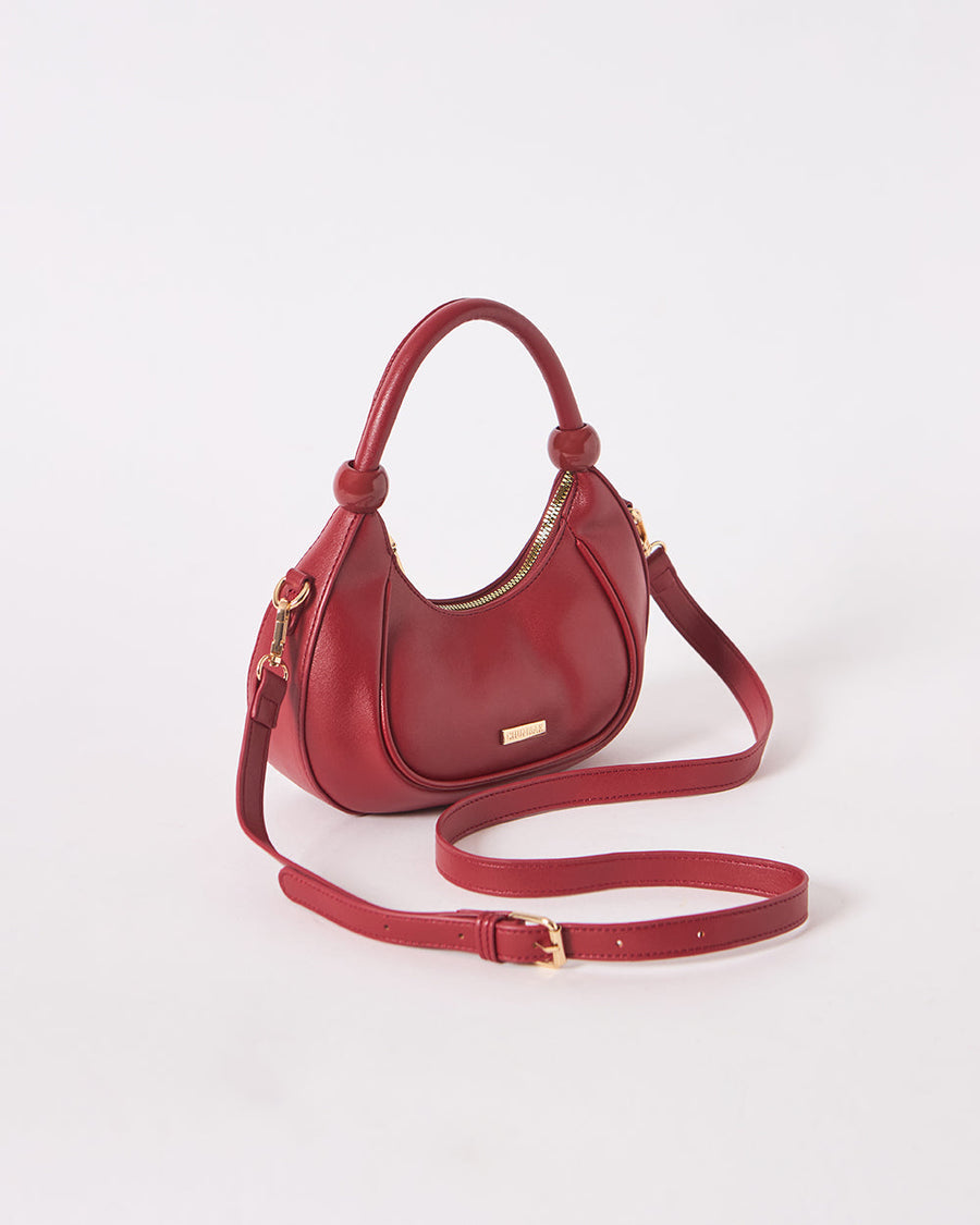 Side angle view of maroon vegan leather crescent handbag highlighting smooth finish and hardware details.