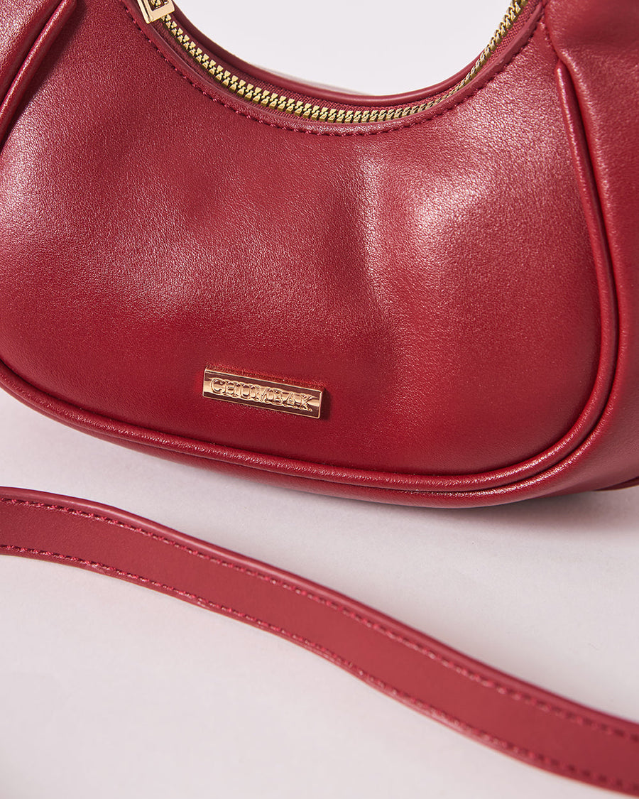 Close-up of the gold branded plaque on the front and smooth texture of the maroon vegan leather crescent handbag.