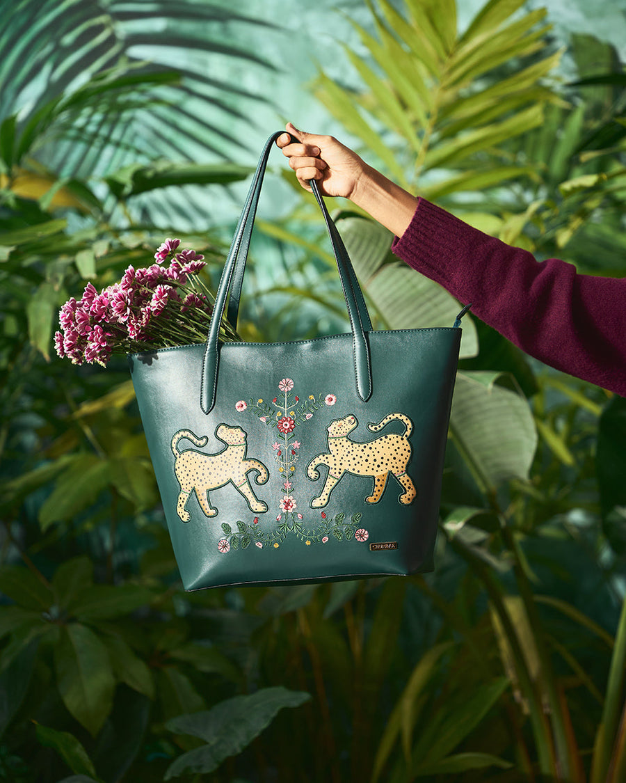 Hand holding deep green vegan leather tote bag decorated with embroidered yellow leopard motifs and floral patterns, filled with pink flowers, outdoor greenery background