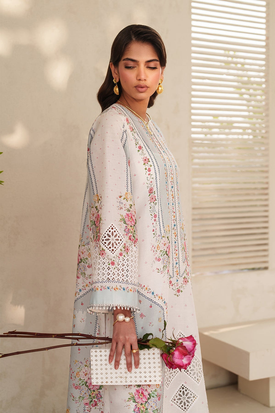 Model holding dupatta draped over shoulders showcasing soft fabric and embroidery details, standing beside plants and textured wall