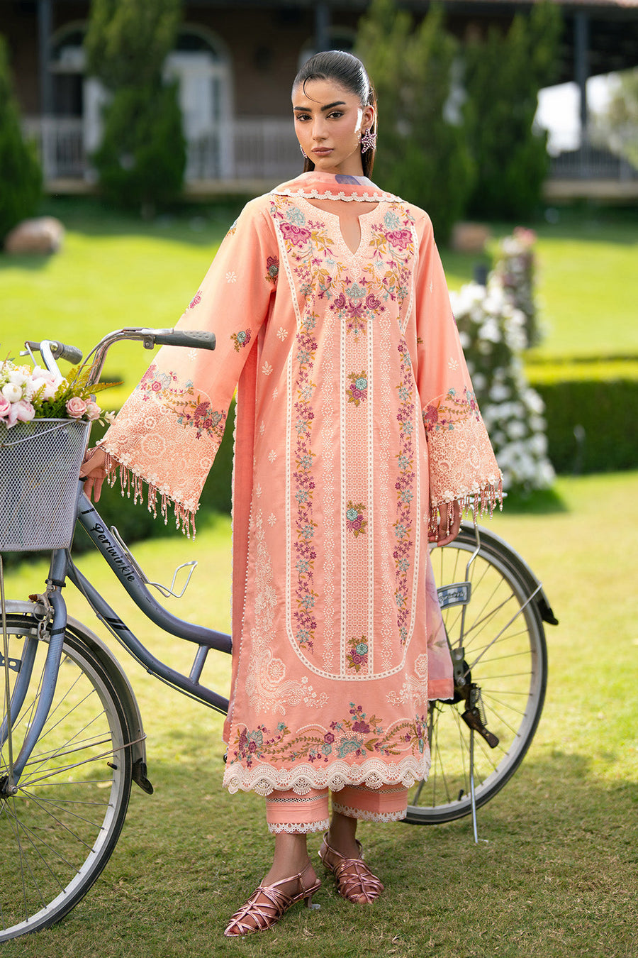 Woman posing next to bicycle wearing peach embroidered lawn suit with layered floral patterns