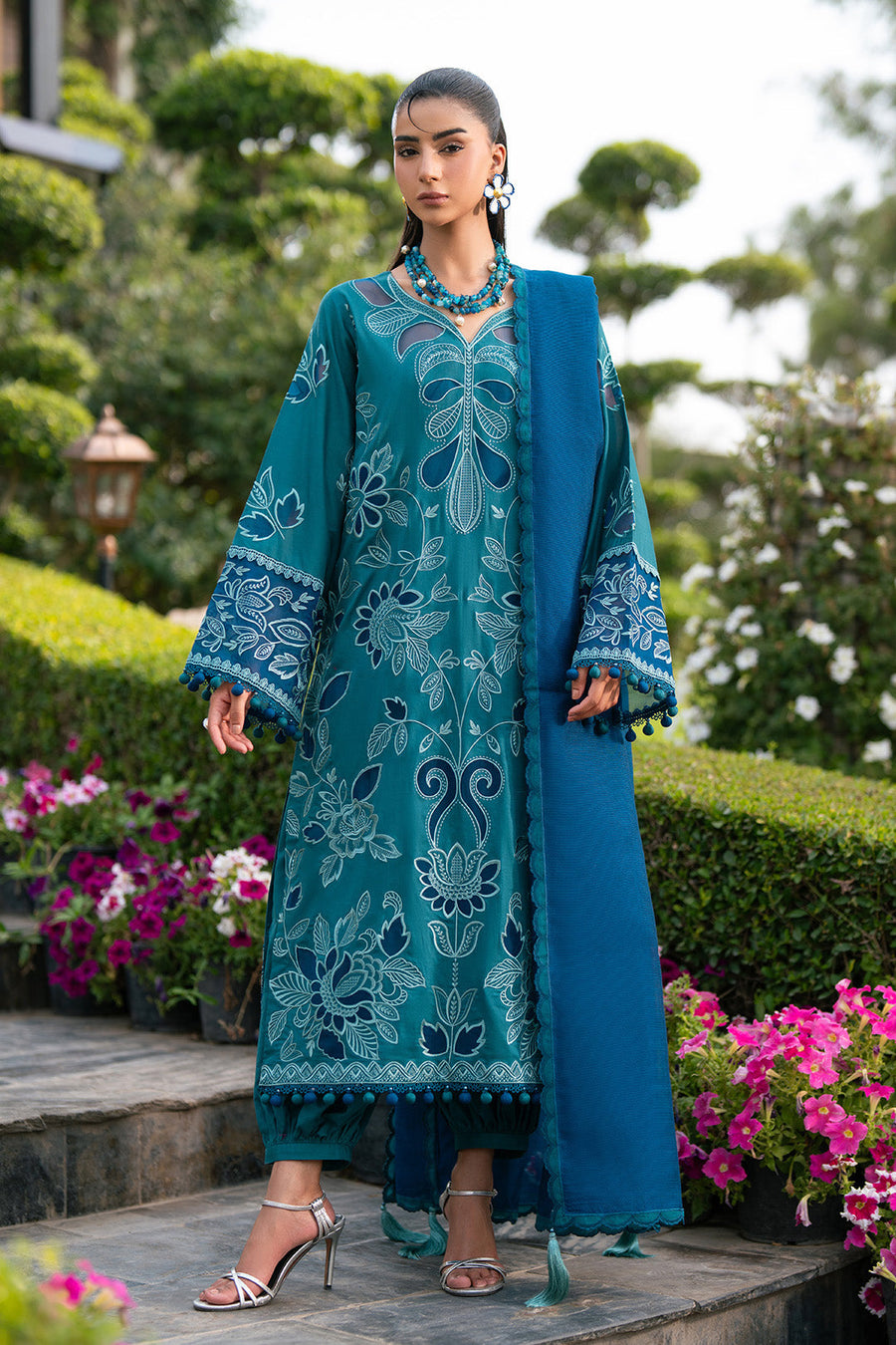 Woman wearing a deep teal-green embroidered lawn suit with pompom trim and matching blue dupatta in garden setting