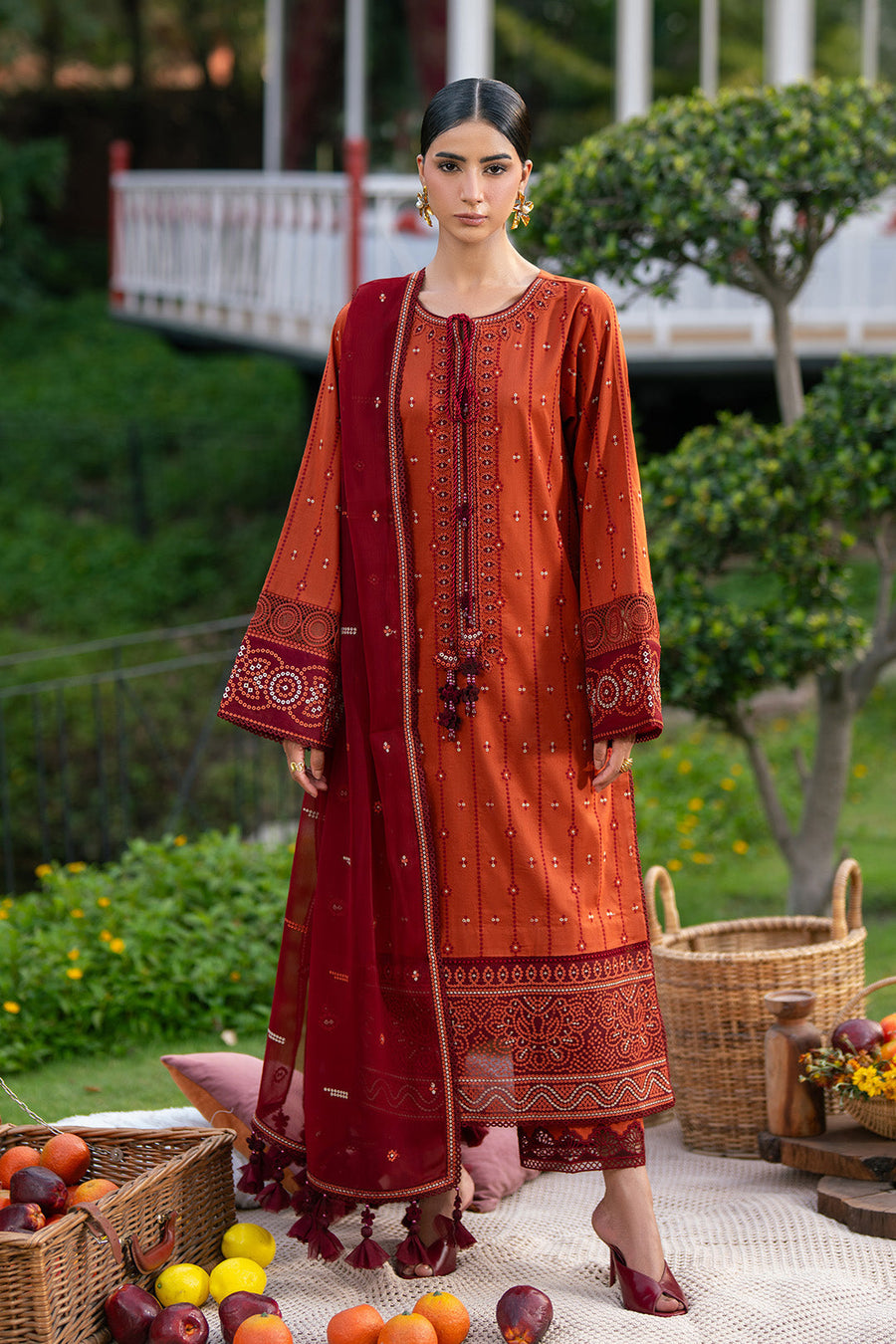 Woman wearing rust orange embroidered lawn suit with maroon dupatta standing outdoors