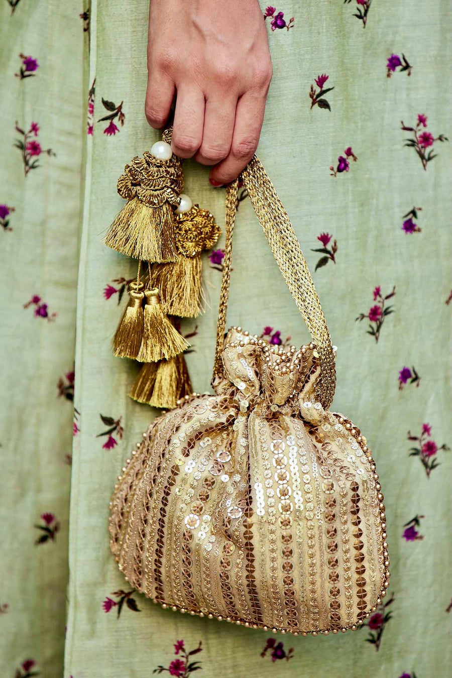 Close-up of hand holding an antique gold embroidered potli bag with tassels against a light green embroidered dress
