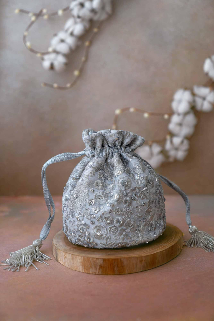 Silver potli bag with intricate floral sequin embroidery and drawstring tassels placed on a wooden round stand with soft decorative background