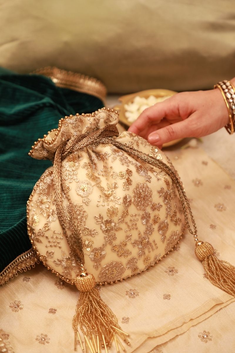 Cream potli bag embellished with gold floral embroidery and beaded tassels, placed on a matching fabric beside a hand adorned with bracelets