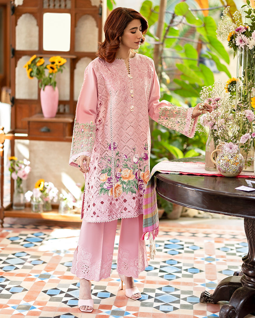 Full front view of a woman in an embroidered pink lawn outfit with floral details, lace trousers, and matching heels, set in a bright floral-decorated room.