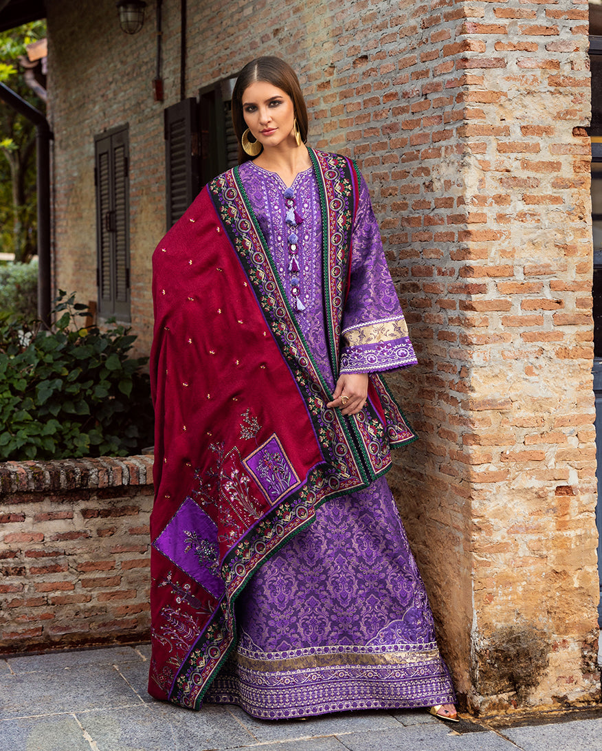 Full-length view of woman draped in a rich crimson shawl with embroidered floral patches and borders over the purple jacquard shirt, standing against a rustic brick wall.