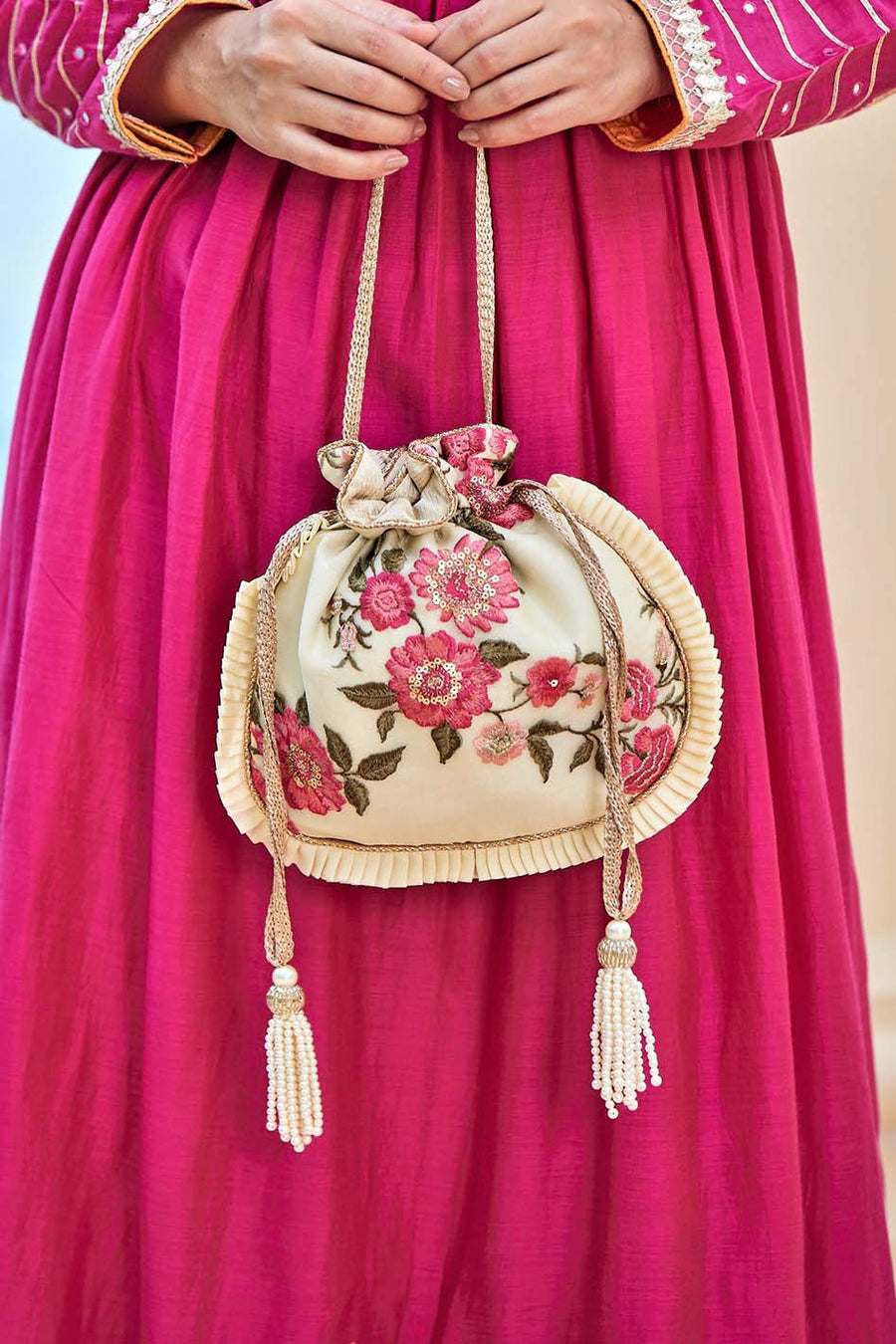 Close-up of a cream embroidered potli bag with pink floral design, pleated edges, and beaded tassels, held by a woman wearing a fuchsia dress.