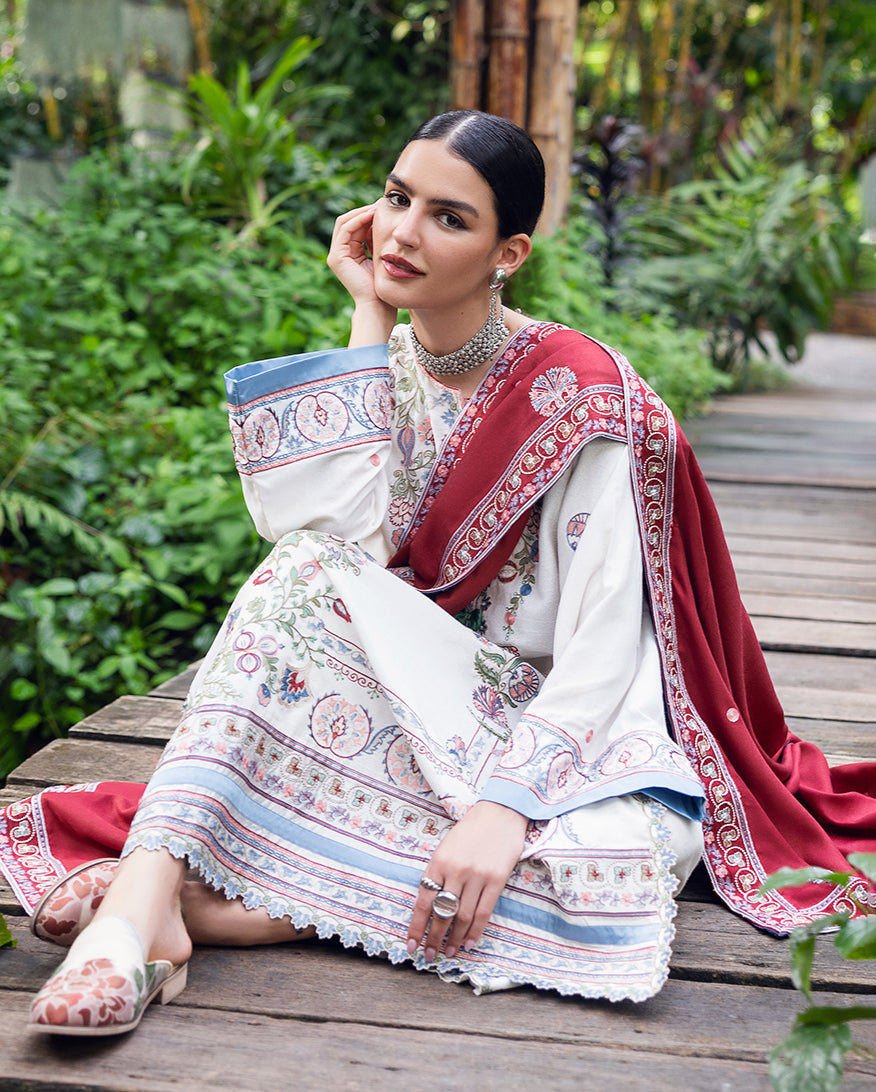 Woman seated on wooden pathway wearing white embroidered Karandi outfit with blue and pink floral accents and deep red shawl, accessorized with rings and choker