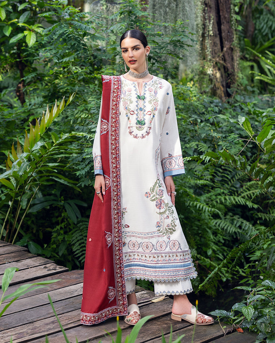 Woman showcasing front view of white Karandi suit with colorful floral embroidery and deep red embroidered shawl, standing on wooden pathway in lush greenery