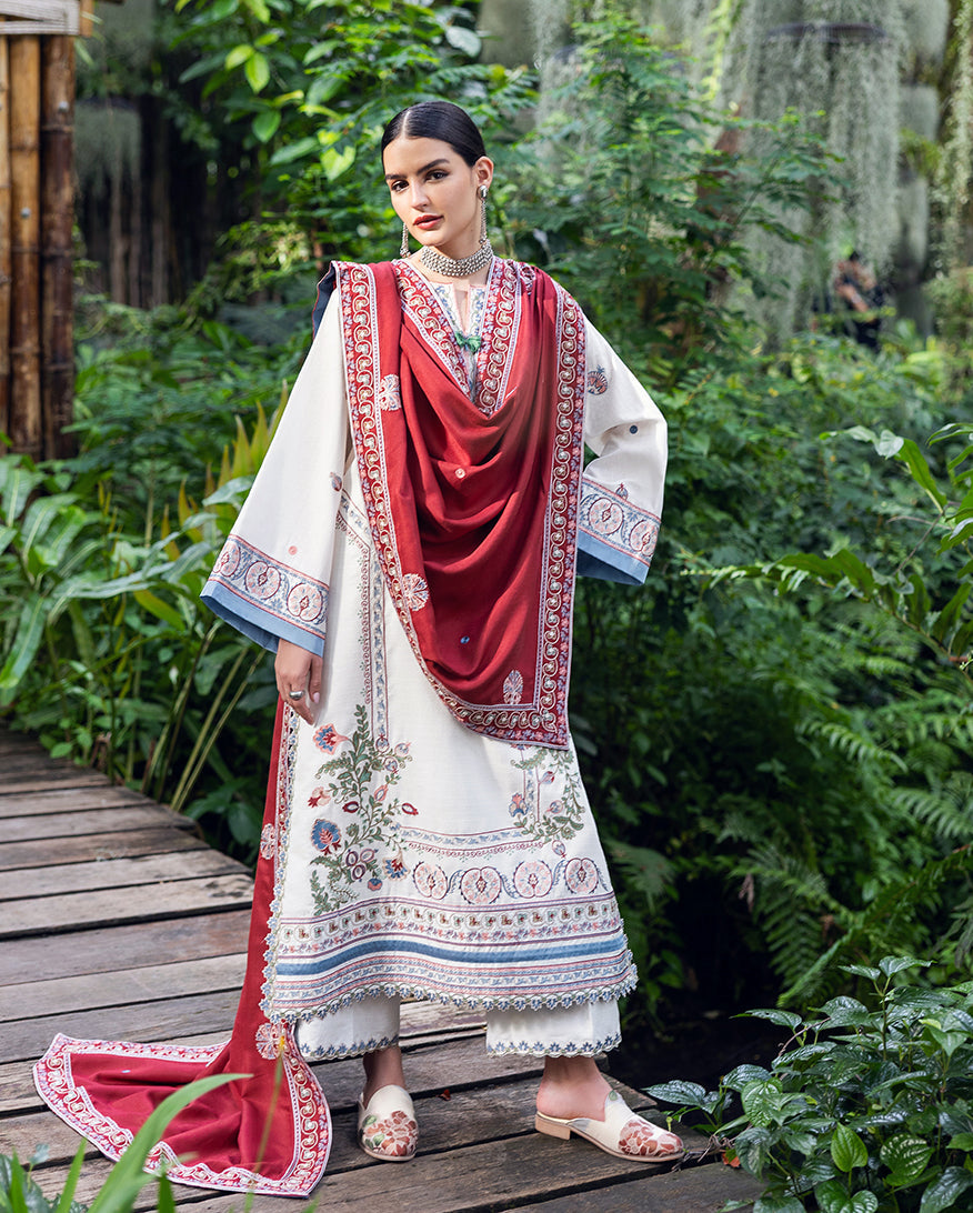 Full length portrait of woman standing outdoors wearing modest white Karandi suit with intricate embroidery and draped vibrant red shawl, paired with slip-on shoes