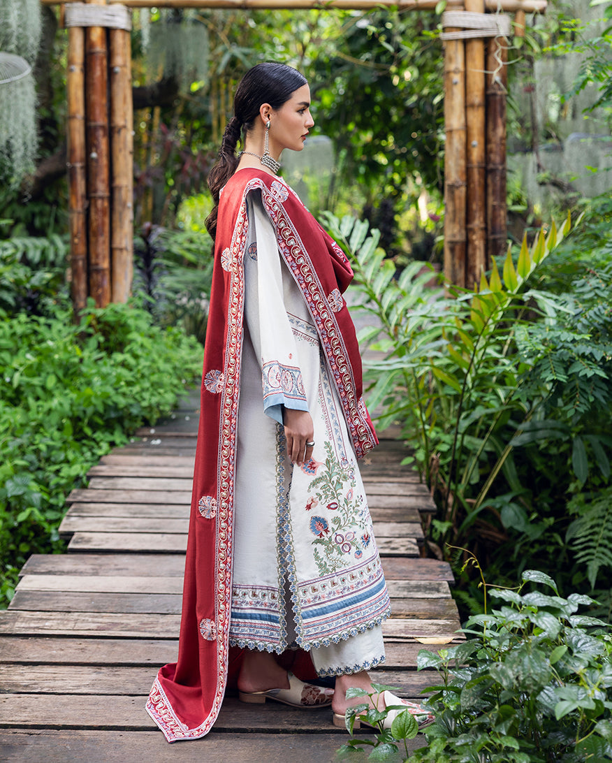 Side profile of woman wearing white embroidered Karandi suit with long sleeves and contrasting red shawl draped over shoulder, outdoors on wooden bridge