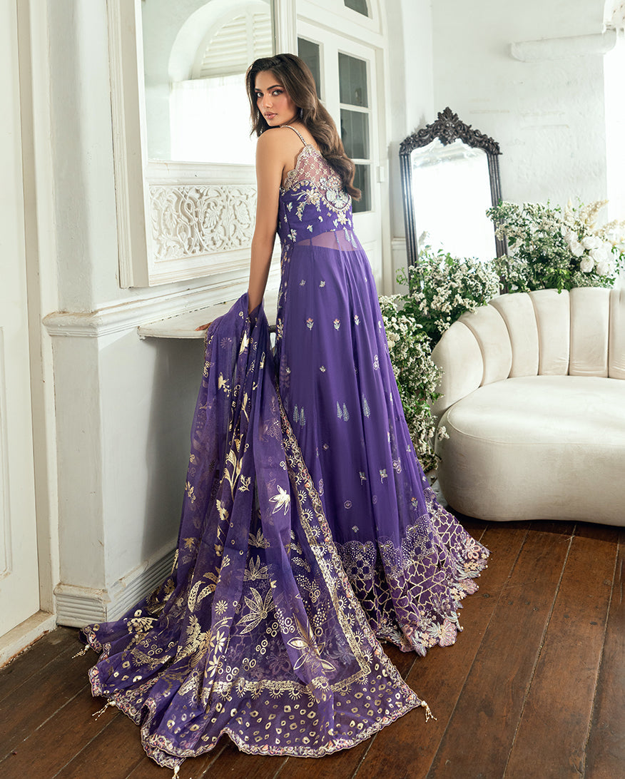 Side back angle of woman seated by a window wearing purple chiffon gown with embroidered details and carrying matching dupatta