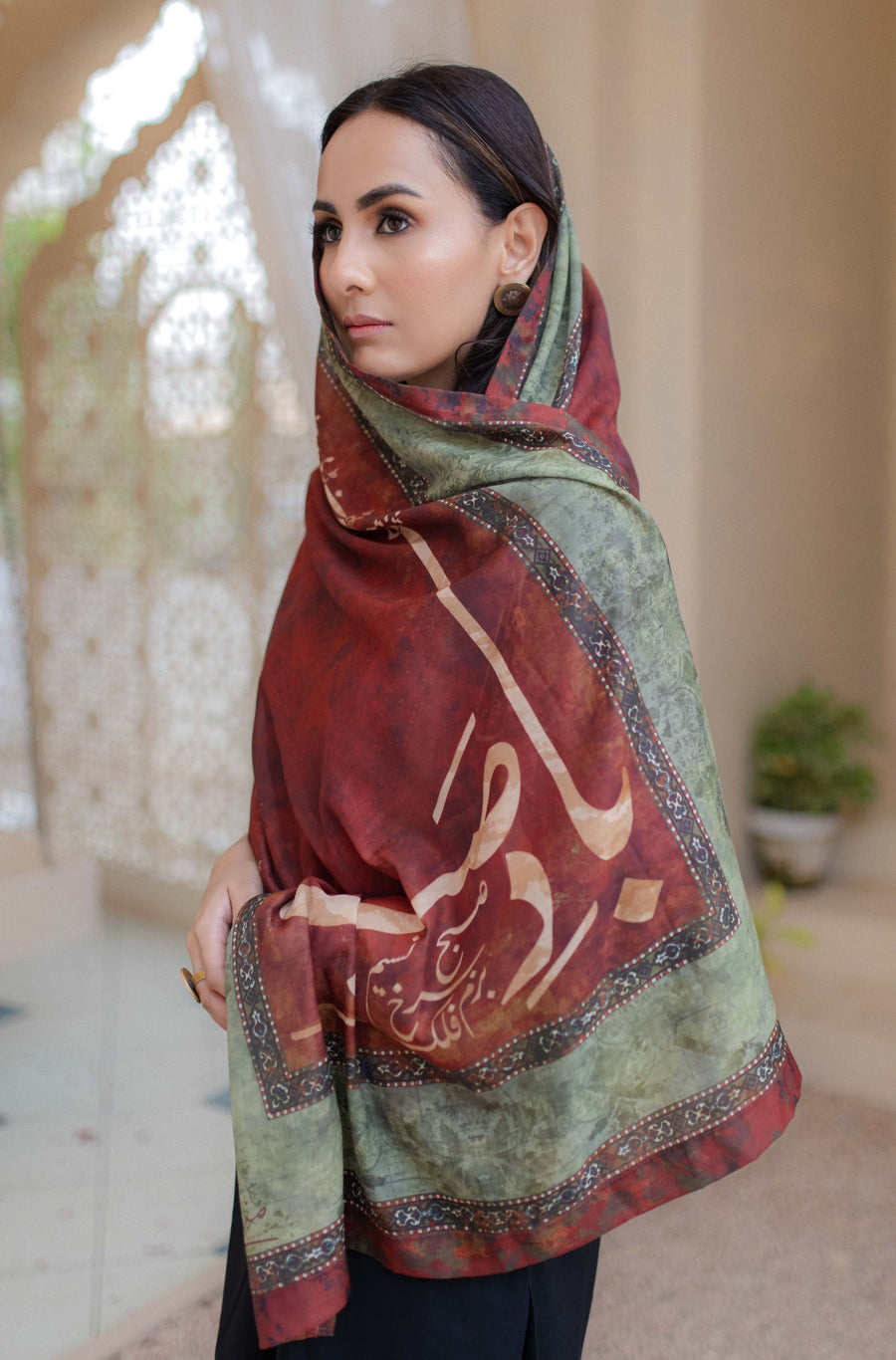 Woman wearing a large rectangular scarf in maroon and muted green with calligraphy and decorative borders, styled modestly over her shoulders.