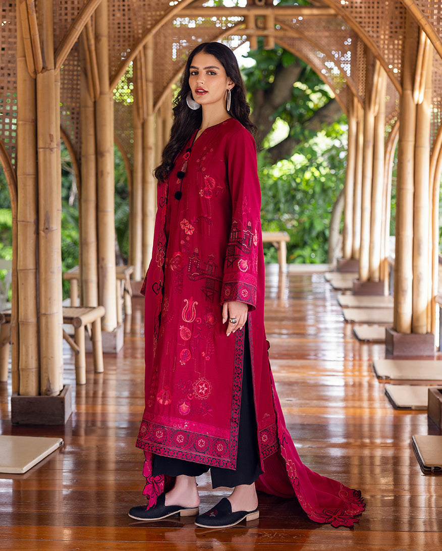 Side view of woman in deep pink embroidered Karandi shirt with black pants and detailed sleeve embroidery, standing in a bamboo structure corridor