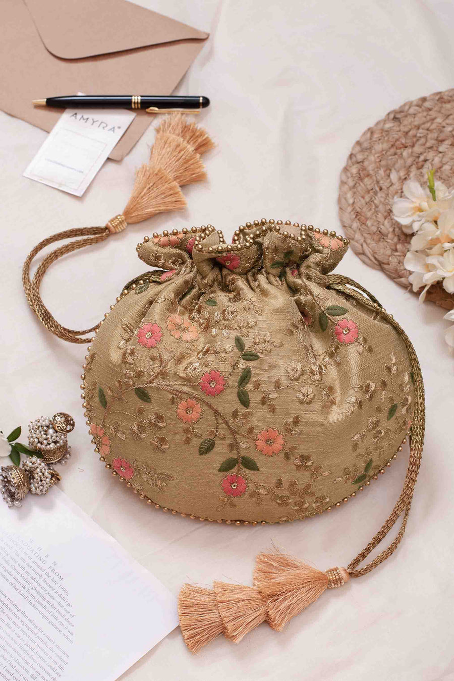 Gold floral embroidered drawstring bag with braided tassel strings laid on fabric with elegant accessories