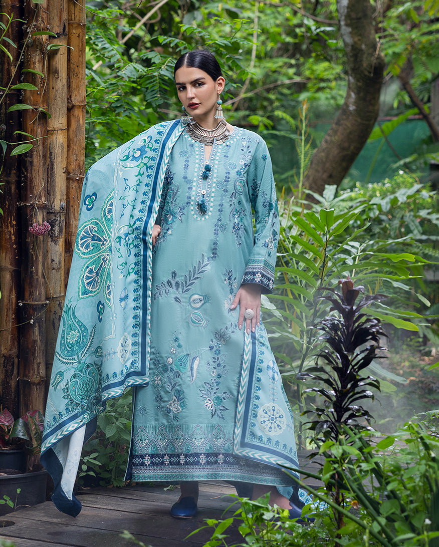 Woman wearing an aqua blue embroidered karandi suit with floral and geometric patterns, holding a matching printed dupatta in a lush green garden setting.