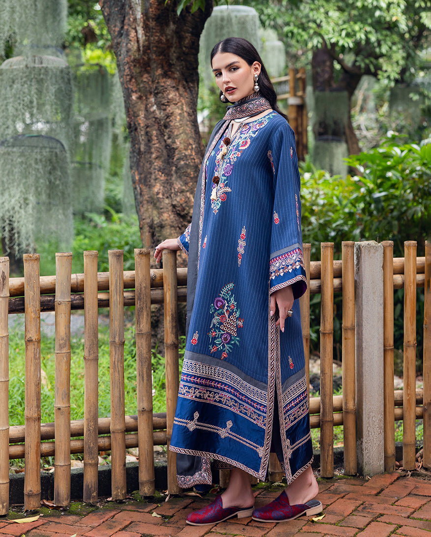 Side view of woman in blue embroidered karandi suit with detailed sleeve borders and grey shawl by bamboo fence