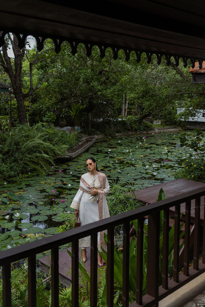 Woman standing on wooden platform next to lily pond, wearing ivory embroidered raw silk suit with detailed dupatta draped elegantly.