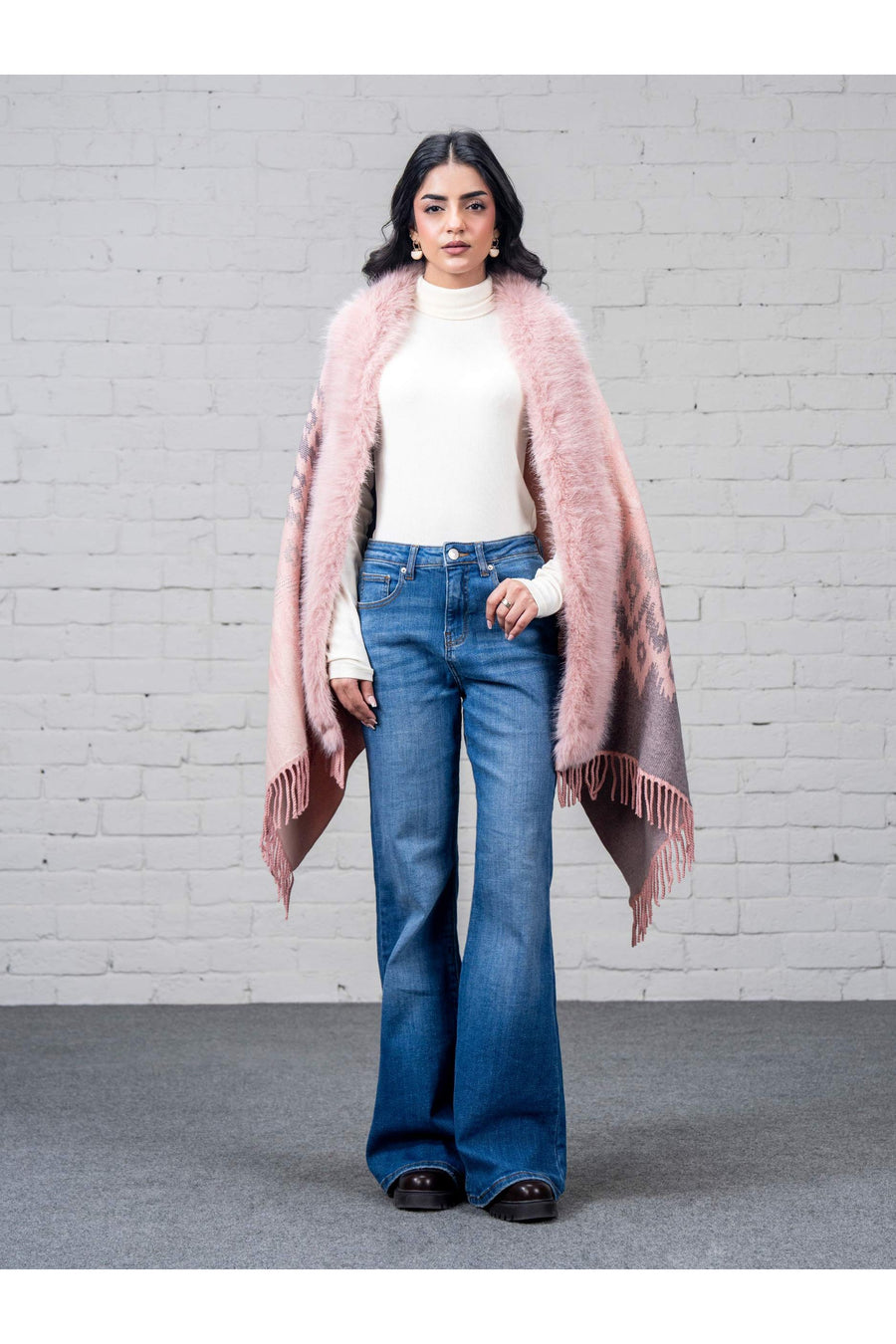 Front view of woman wearing pink wool cape with faux fur trim, white turtleneck, and blue flare jeans against brick wall