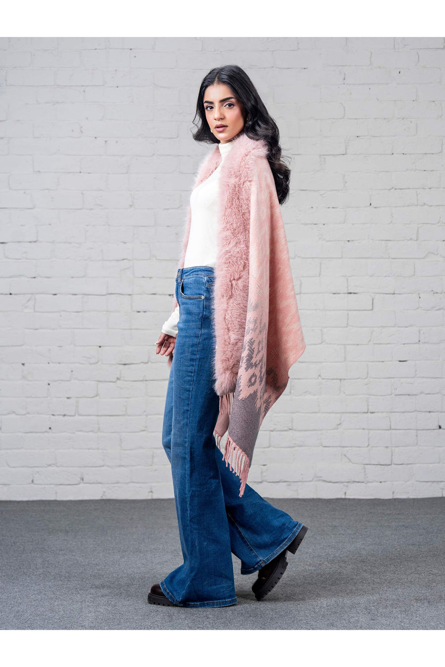 Side view of woman modeling pink wool cape with patterned details and faux fur trim, paired with blue flare jeans and black shoes