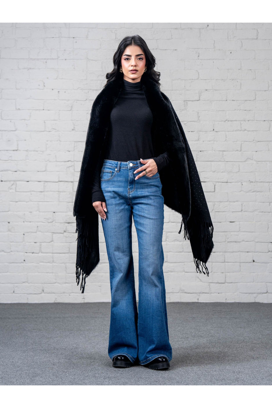 Woman wearing black wool cape shawl with faux fur trim, paired with black turtleneck and blue flared jeans, standing facing forward